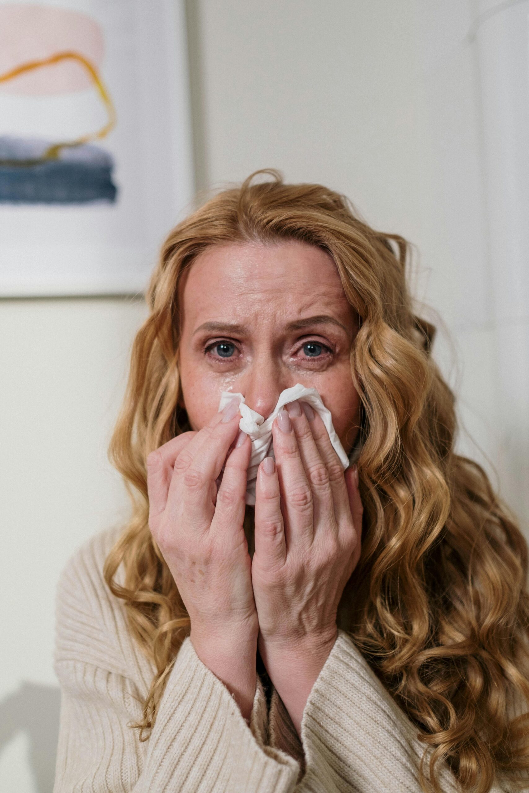 Blonde woman with curly hair sneezing indoors, using tissue for a runny nose.