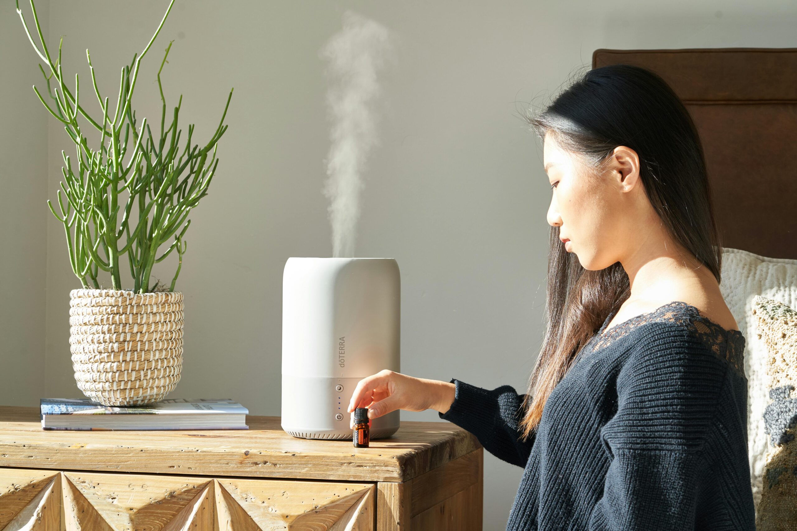Asian woman uses essential oil diffuser on wooden table beside green plant in cozy room.