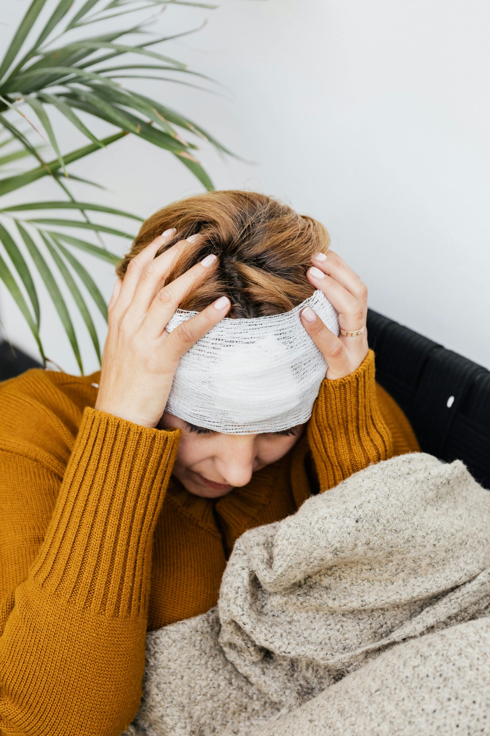 Woman with a bandage holding her head, expressing pain, indoors near a potted plant.