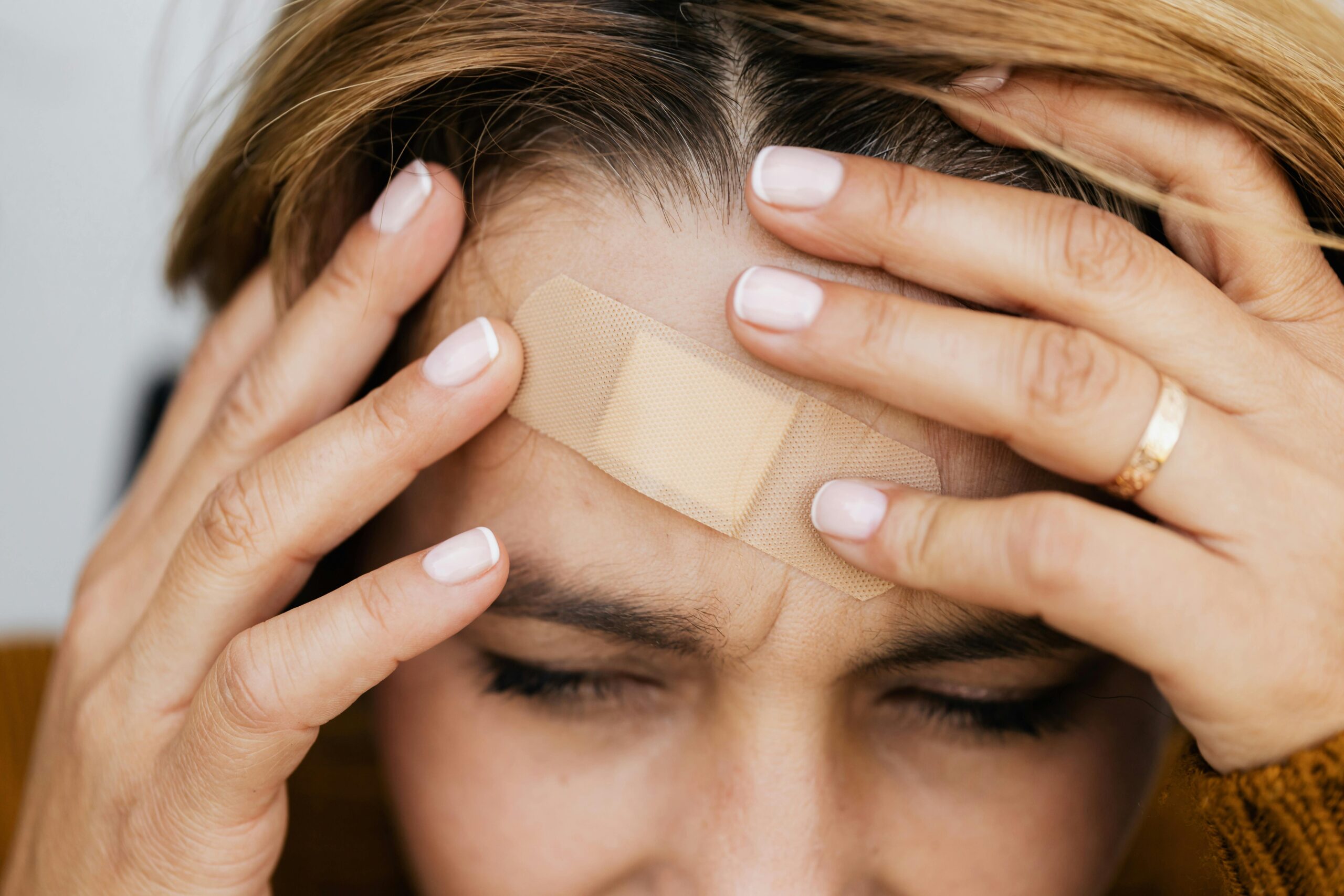 A person with a band aid on their forehead holds their head in pain, eyes closed.