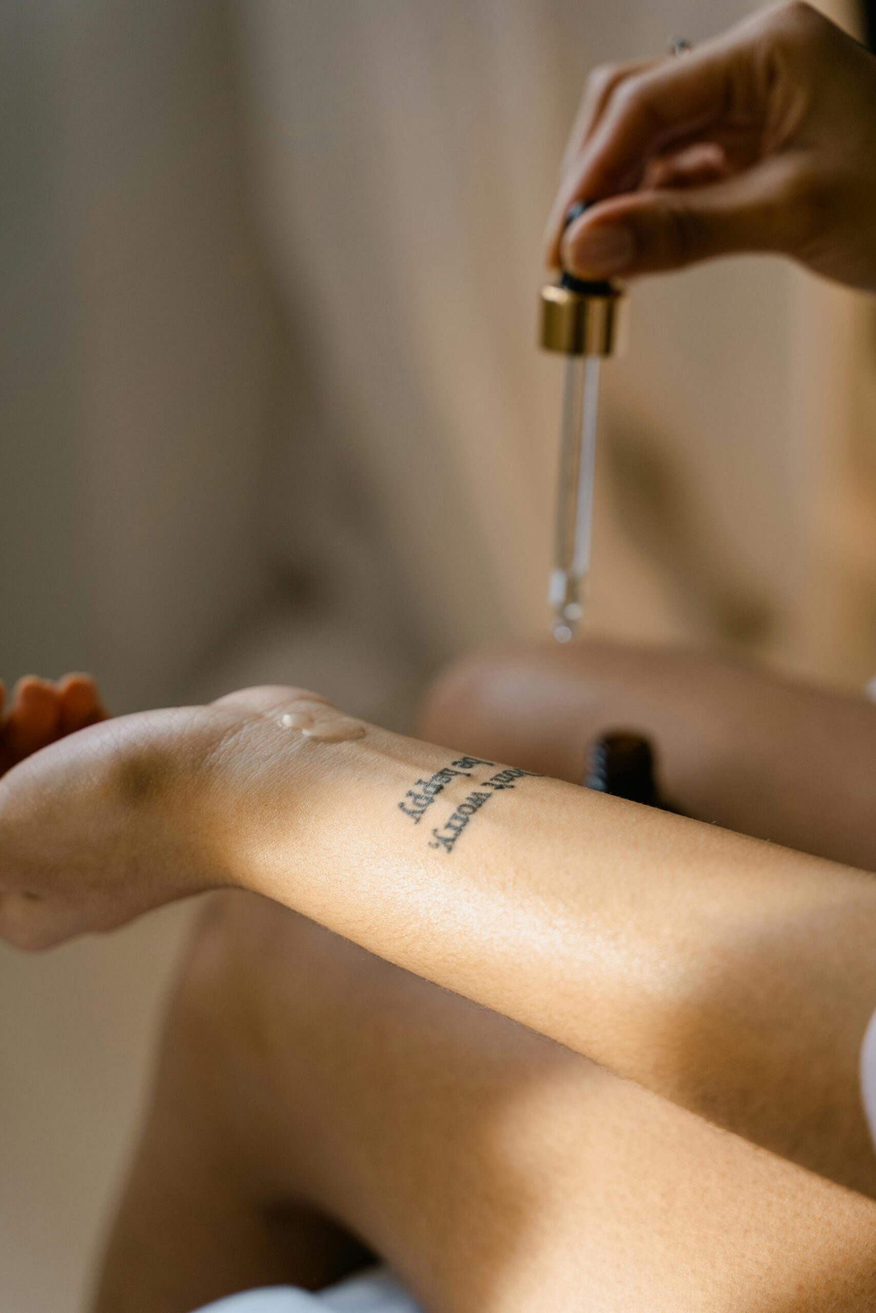 A close-up of a tattooed arm with a dropper applying skin care oil.