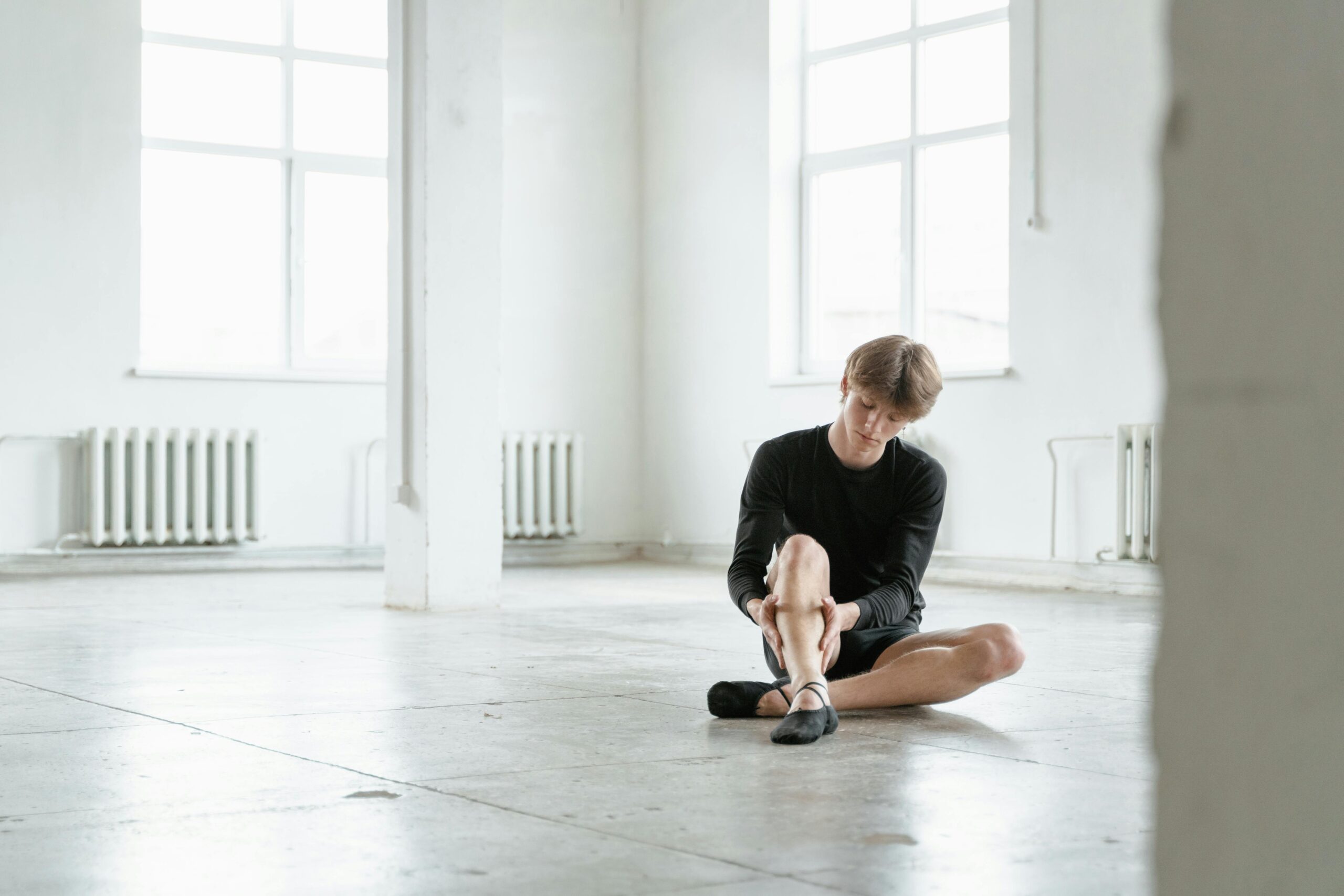 A young dancer in black attire sitting and massaging leg in a bright, spacious studio.