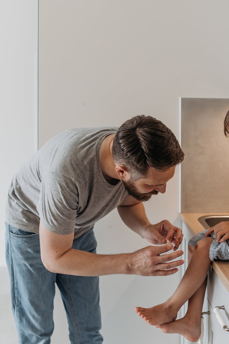 A father is applying a bandage to his son's knee as they sit on a kitchen countertop.