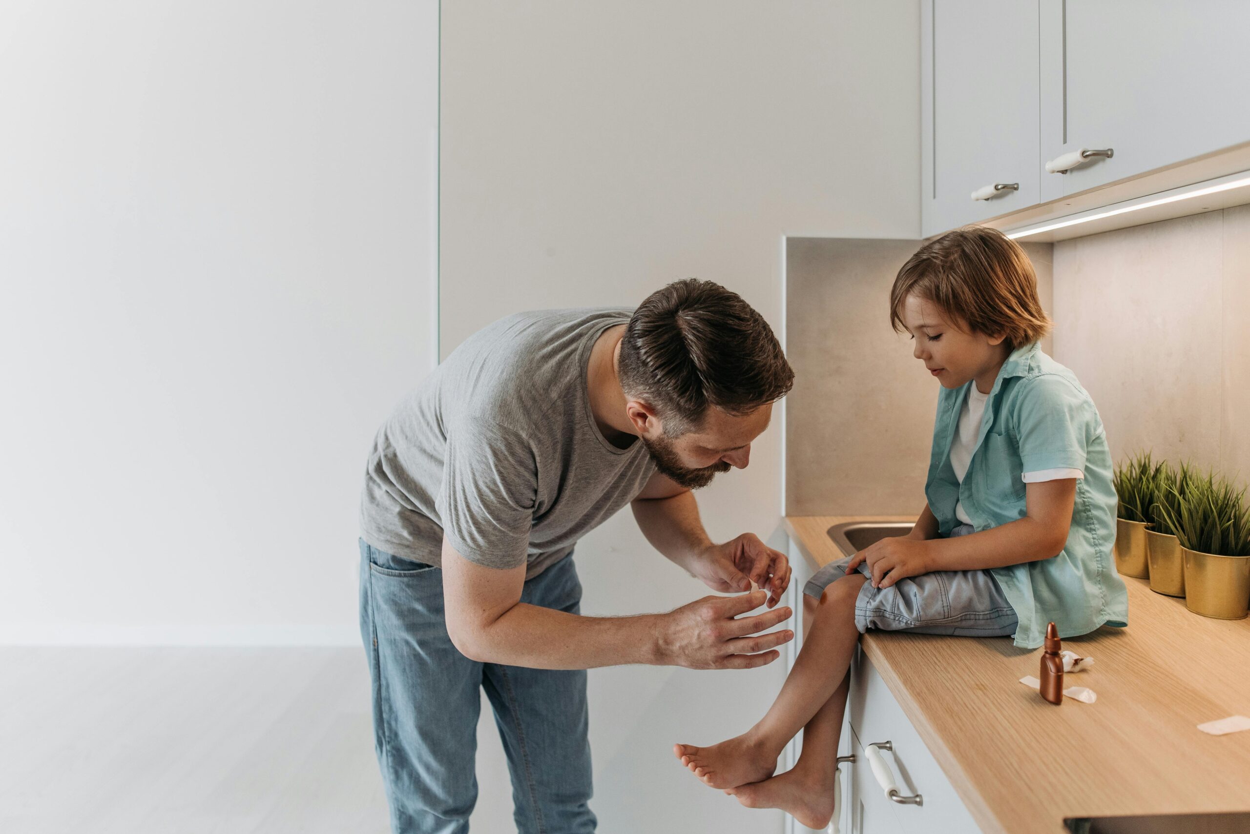 A father is applying a bandage to his son's knee as they sit on a kitchen countertop.