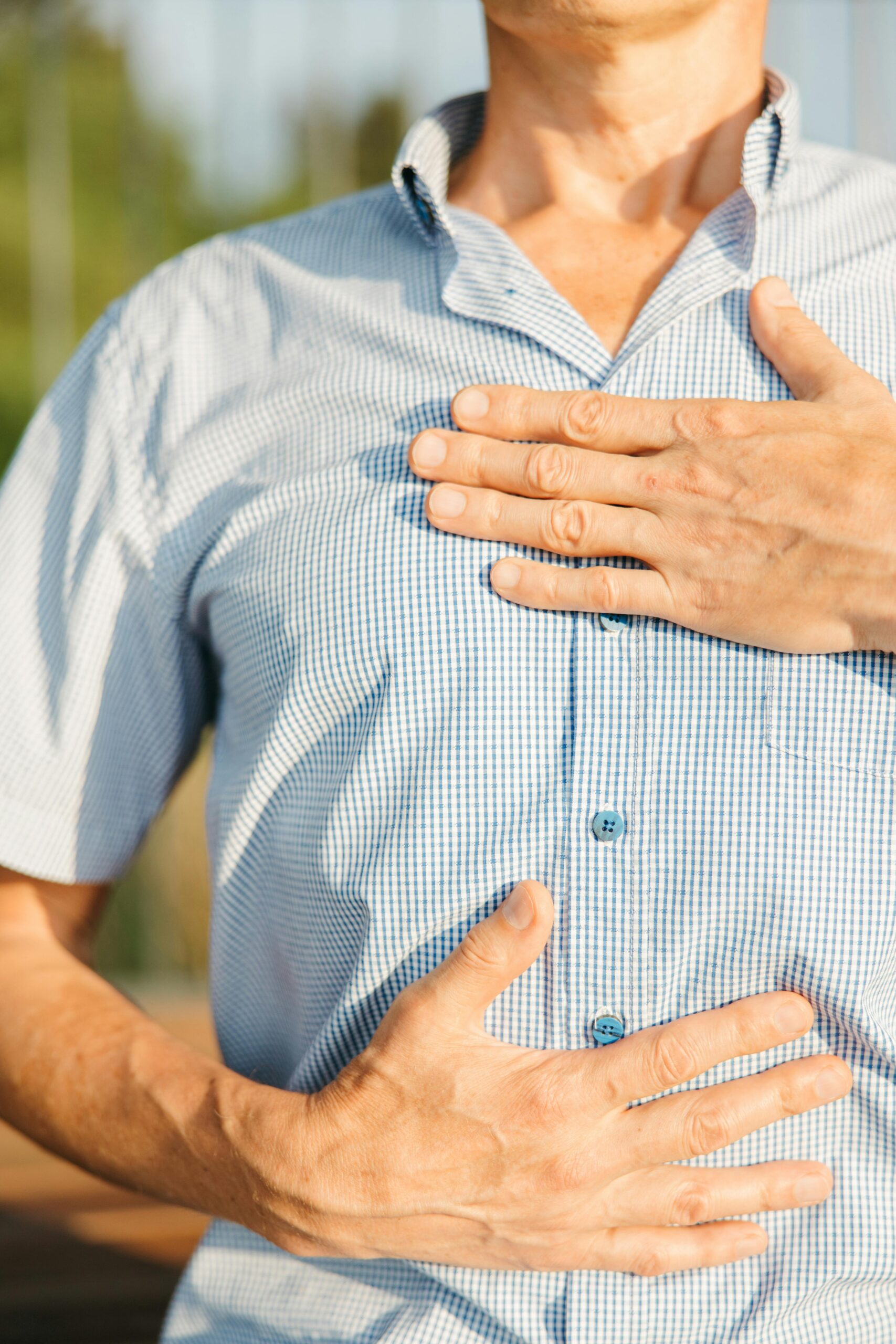 Close-up of a man in a blue shirt holding his chest and stomach outdoors, indicating discomfort.