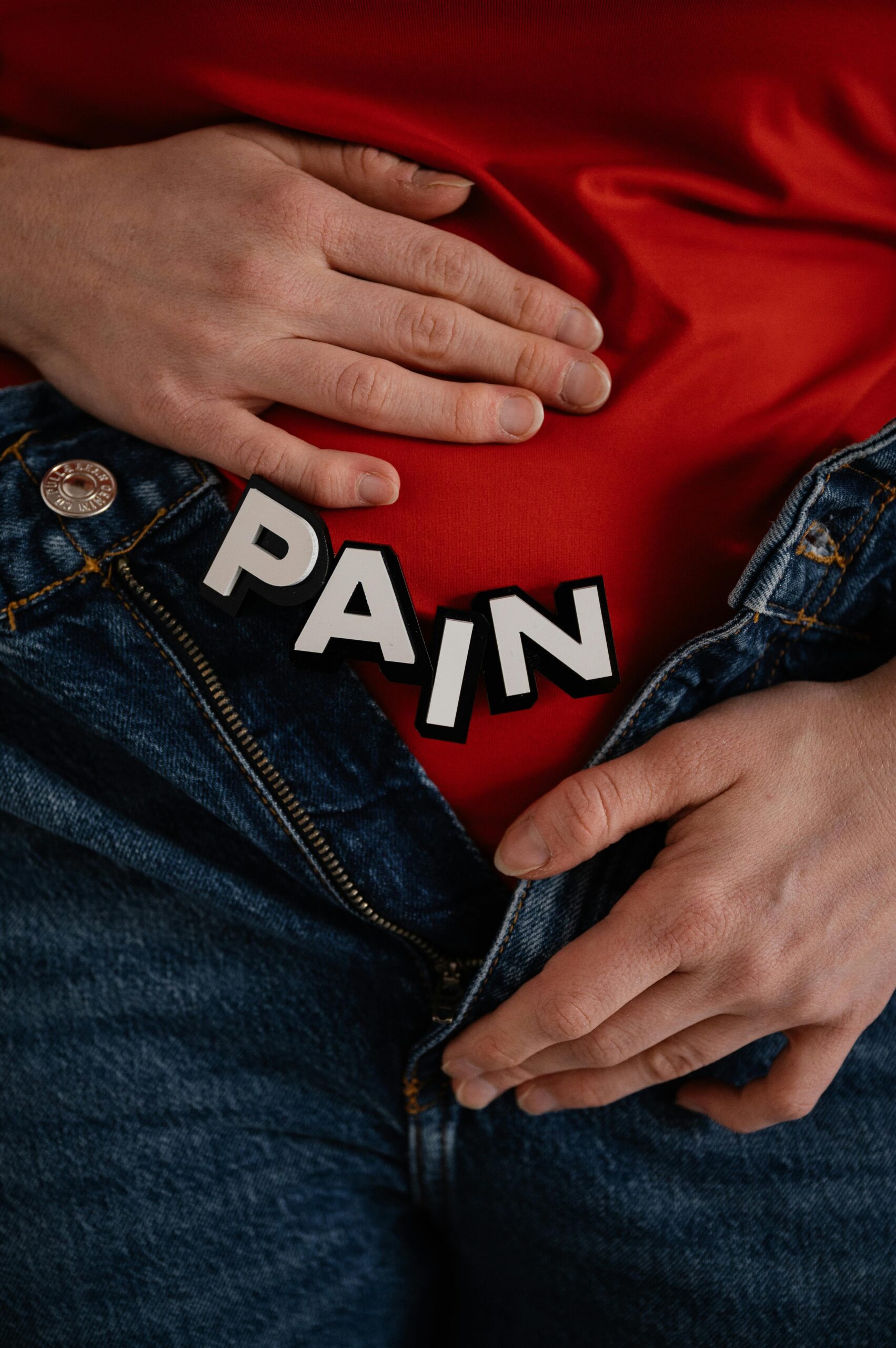 Close-up of a person in jeans holding their stomach with 'PAIN' text overlay.