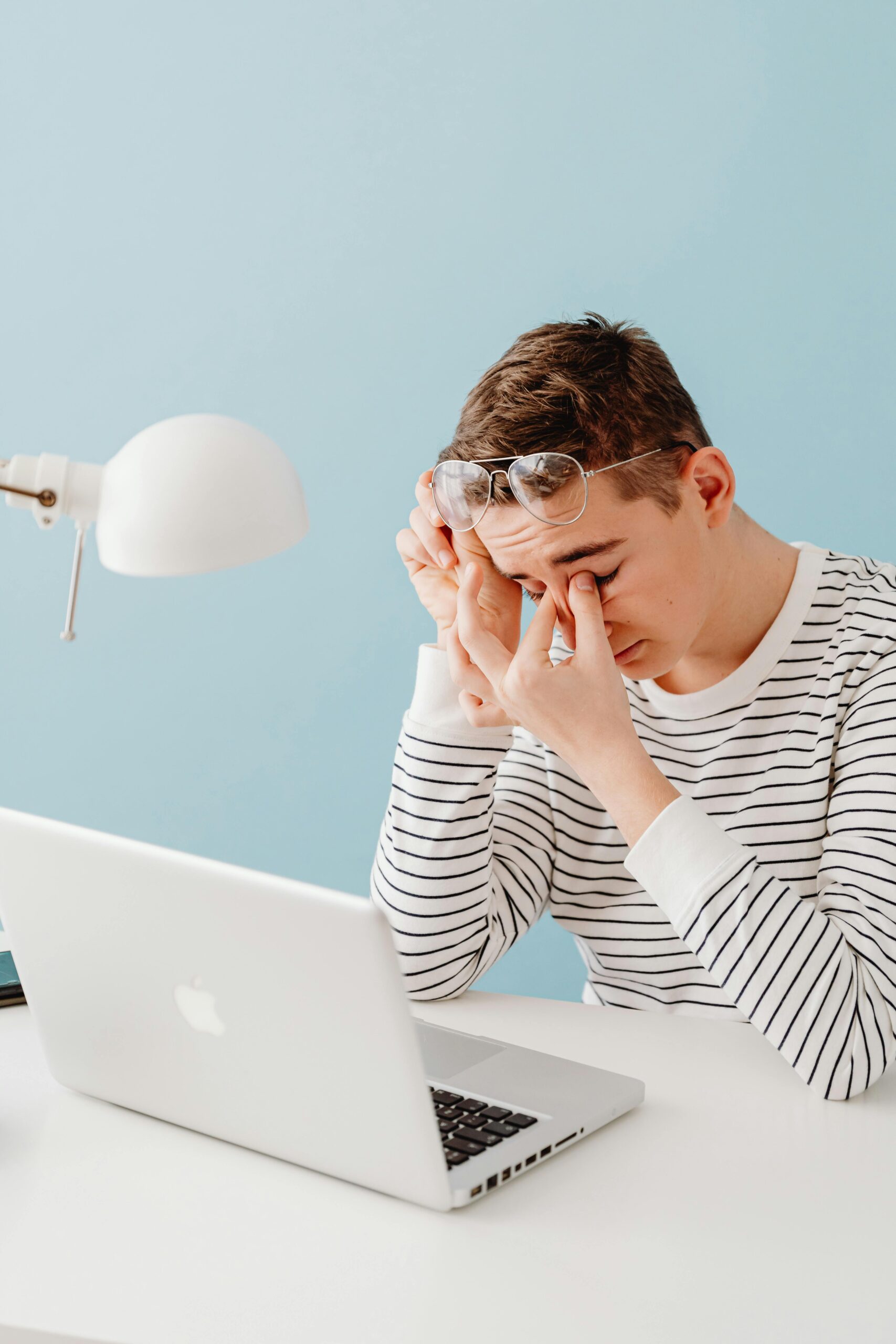 Teenager in striped shirt showing fatigue while sitting at a desk with a laptop and lamp.
