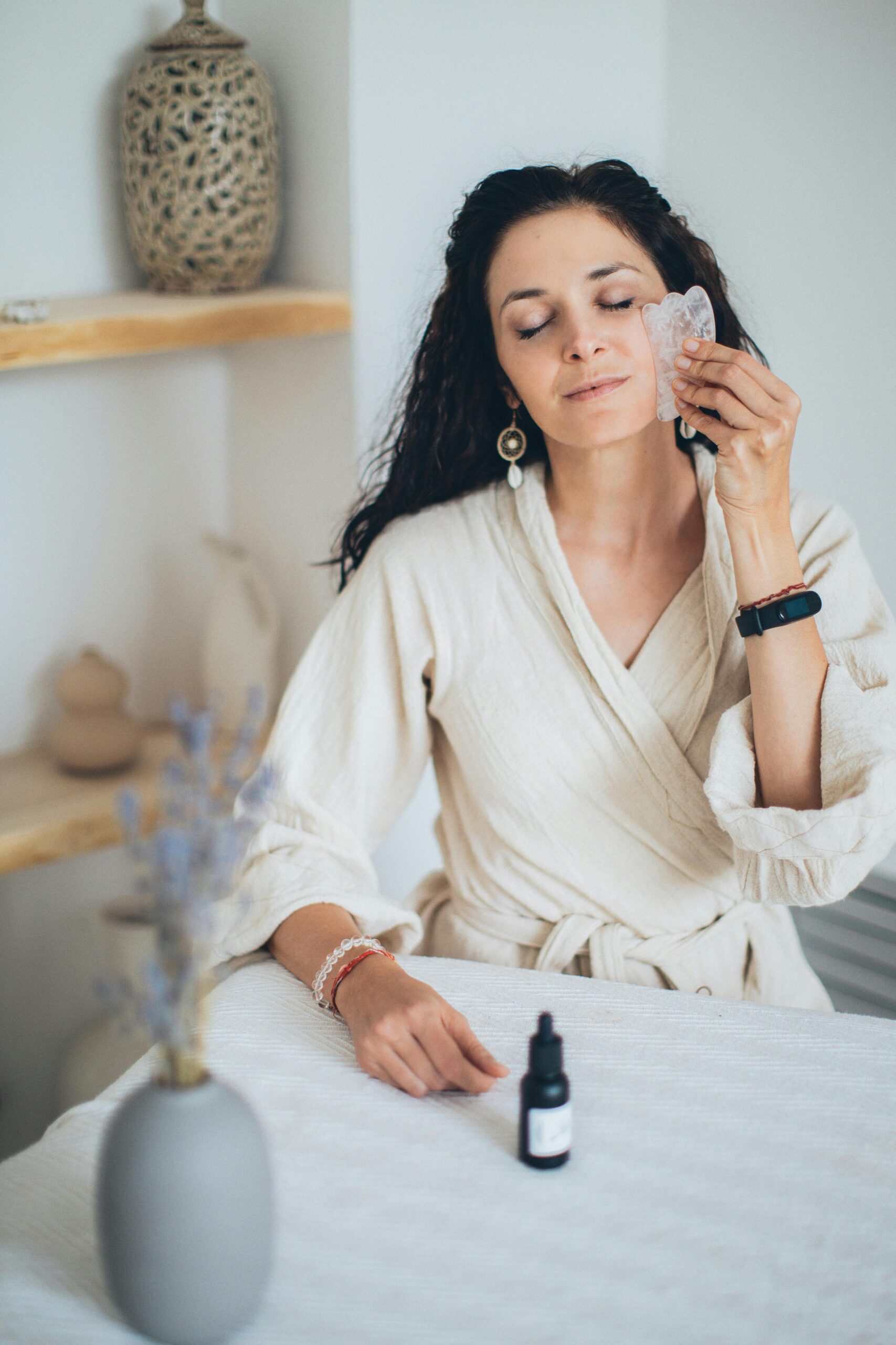 A woman in a bathrobe using a gua sha tool for a relaxing facial massage at home.