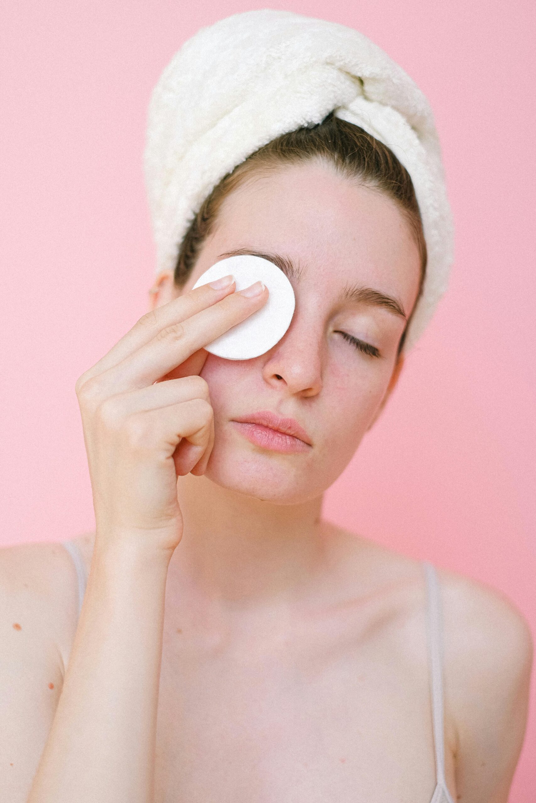 Young woman cleansing face with cotton pad on pink background