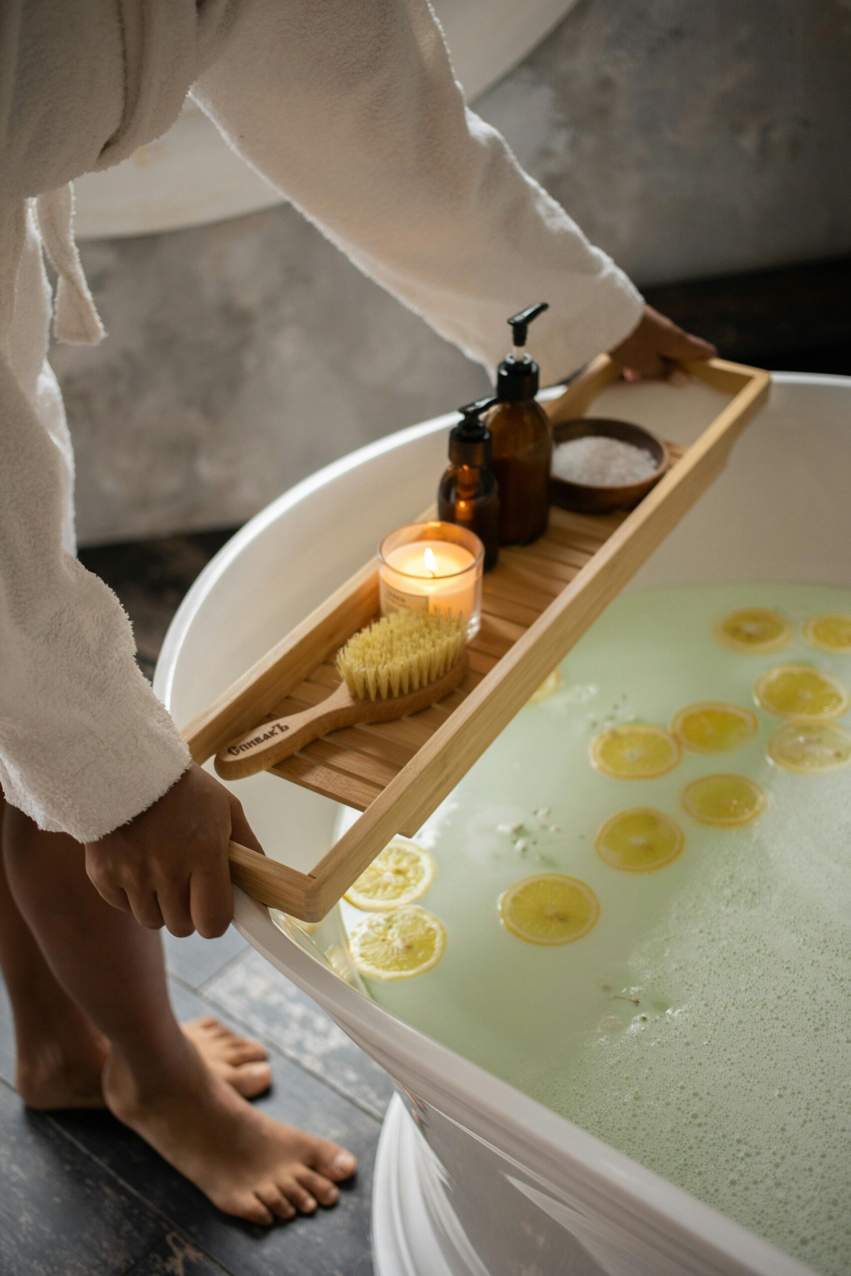 Unrecognizable barefoot female in bathrobe putting wooden tray with spa supplies on tub with lemon slices on water surface during skincare routine