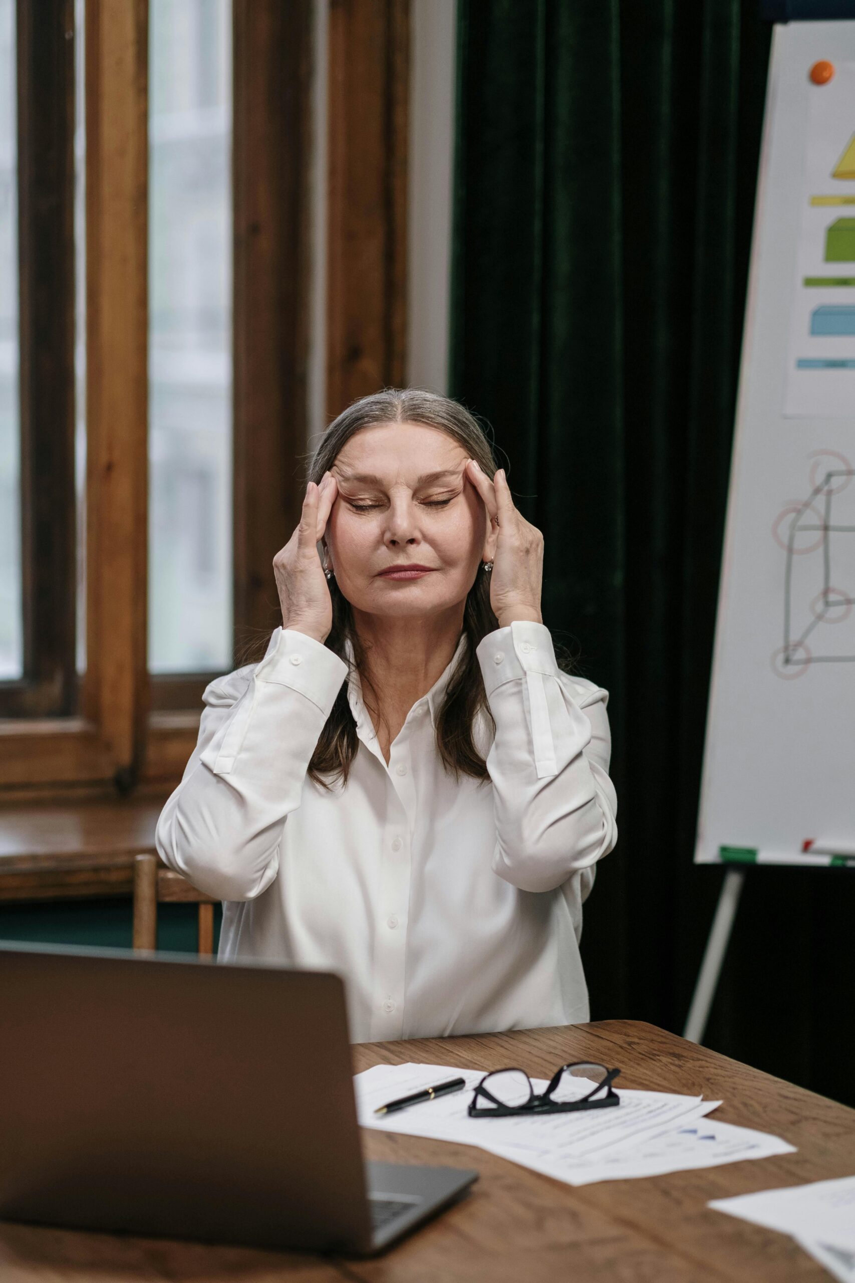 Elderly woman in white shirt massaging temples at desk, laptop open.