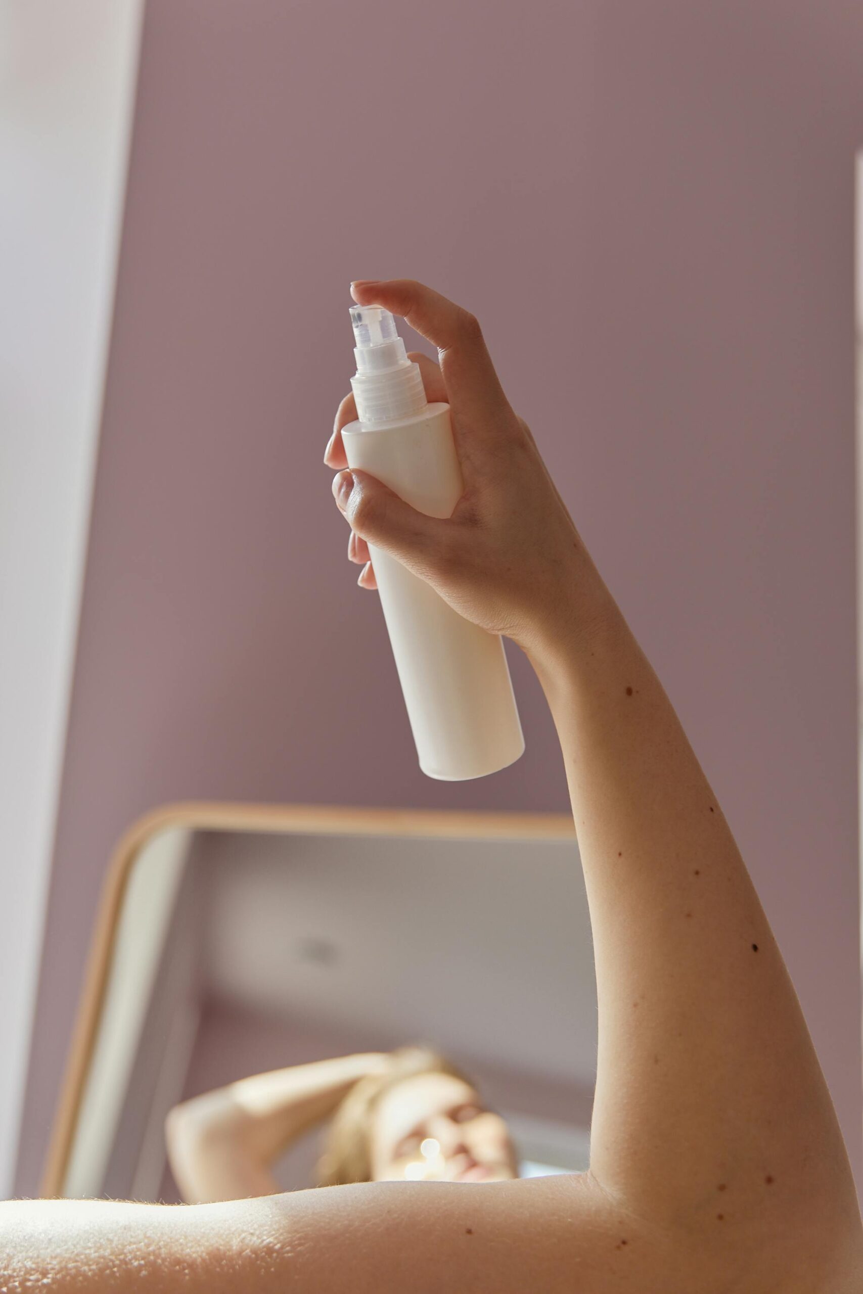 A hand holding a white spray bottle, positioned in front of a mirror, with soft lighting.